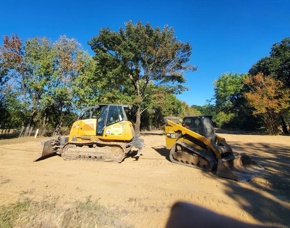 Two yellow bulldozers sit parked on a cleared patch of dirt, backed by trees and a bright blue sky.